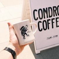 a person holding a coffee mug in front of a sign that says condor coffee